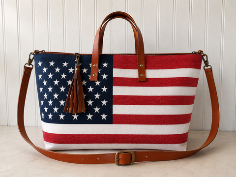 A tote bag featuring a woven American flag pattern with red and white stripes and a navy blue field with white stars, finished with brown leather handles, a matching shoulder strap, and a brown leather tassel charm.